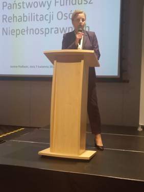 Pokaż zdjęcie: Sala konferencyjna, przy drewnianej mównicy stoi kobieta, blondynka z mikrofonem w ręku, ubrana w ciemny garnitur. W tle wyświetlany jest slajd z prezentacji