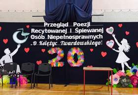 Scena przygotowana na XX Przegląd Poezji Recytowanej i Śpiewanej Osób Niepełnosprawnych „O Księżyc Twardowskiego”. Na czarnym tle widnieje nazwa wydarzenia, kolorowa liczba 20, czerwone serca oraz papierowe sylwetki i kwiaty. Przed dekoracją ustawiono krzesła i stół.