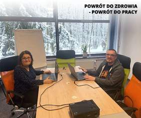 Pokaż zdjęcie: Kobieta i mężczyzna siedzą przy dużym biurku, na stole dwa laptopy, za nimi tablica. W tle zaśnieżony las.