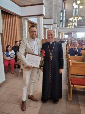 Pokaż zdjęcie: W kościele stoi obok siebie dwóch panów. Jeden z nich jest osobą duchowną, a drugi świecką ubraną w garnitur. W tle widać ławki i siedzących na nich ludzi. 