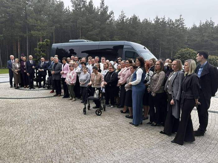 Pokaż zdjęcie: Grupa kilkudziesięciu osób stoi przed autobusem zaparkowanym na placu wyłożonym kostką brukową. W tle znajduje się las iglasty. 