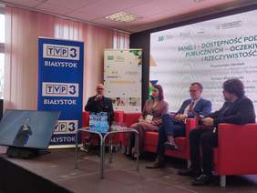 Pokaż zdjęcie: Zdjęcie przedstawia panel dyskusyjny podczas konferencji ZUS. Na scenie czterech panelistów siedzi na czerwonych fotelach. Mężczyzna po lewej, w okularach, mówi do mikrofonu, a pozostali – kobieta w bordowym garniturze oraz dwaj mężczyźni – słuchają z uwagą. W tle widoczny jest ekran z tematem panelu, banery TVP3 Białystok i ZUS oraz monitor z tłumaczem języka migowego. Na stoliku przed panelistami stoją butelki wody i szklanki.