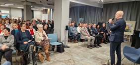 Pokaż zdjęcie: Mężczyzna na środku sali przemawia do mikrofonu. Widoczny publiczność - liczni uczestnicy słuchają wystąpienia.