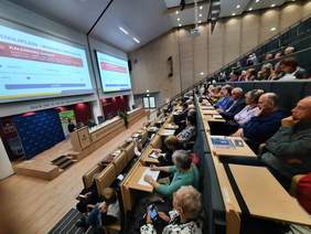 Pokaż zdjęcie: W sali wykładowej Politechniki Łódzkiej siedzą na krzesłach seniorzy. Słuchają wykładu i patrzą na prelegentkę. 