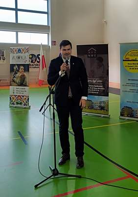 Pokaż zdjęcie: Młody mężczyzna w ciemnym garniturze przemawia do mikrofonu, jest w Sali gimnastycznej. Za nim banery reklamowe.
