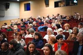 Pokaż zdjęcie: Zdjęcie z widowni teatru lub kina. Widać rzędy czerwonych foteli, w których siedzi duża grupa osób w różnym wieku, skierowanych przodem do niewidocznej sceny. W prawym górnym rogu znajduje się fragment drewnianej ściany z kwadratowym oknem.