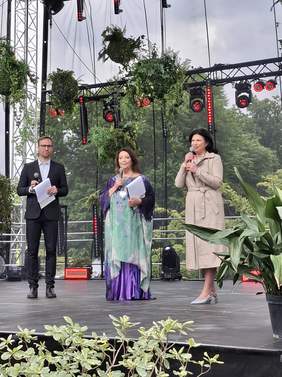Pokaż zdjęcie: Scena w ogrodzie botanicznym otoczona kwiatami i roślinami. Na scenie mężczyzna i dwie kobiety przemawiają do mikrofonów.