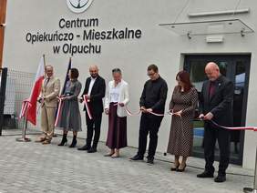 Pokaż zdjęcie: Grupa siedmiu osób stoi przed wejściem do Centrum Opiekuńczo-Mieszkalnego w Olpuchu i wspólnie przecina biało-czerwoną wstęgę. Obok znajduje się flaga Polski.