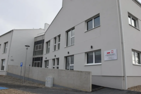 Pokaż zdjęcie: Nowoczesny, jasny budynek z dwoma kondygnacjami, dużymi oknami i łącznikiem przeszklonym; przed nim chodnik, znak parkingu i niski betonowy mur, na ścianie tablica z godłem Polski.  
