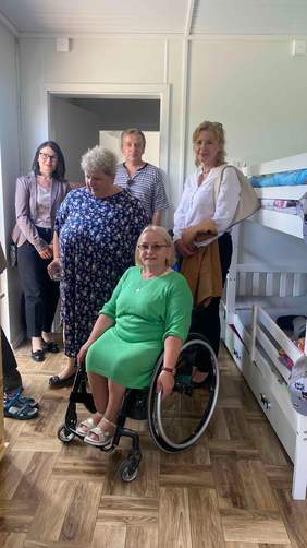 Pokaż zdjęcie: wewnątrz kontenera mieszkalnego pięć osób w tym jedna kobieta na wózku inwalidzkim zwiedza pomieszczenia mieszkalne