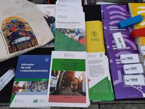 Pokaż zdjęcie: Na stoliku leżą różne materiały promocyjne i reklamowe. Są to: informatory, katalogi, poradniki, ulotki, długopisy i torby. 