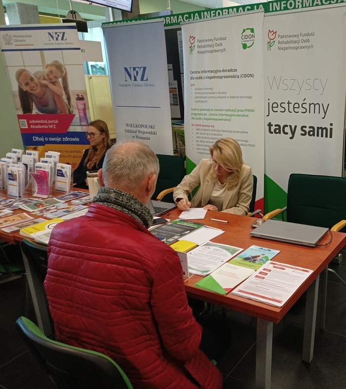 Pokaż zdjęcie: Kobieta reprezentująca Oddział Wielkopolski PFRON udziela informacji starszemu mężczyźnie z niepełnosprawnością narządu słuchu, pisząc na kartce odpowiedź. Na stoliku Oddziału ułożone są różne ulotki, w tle znajdują się dwa banery: PFRON oraz CIDON.