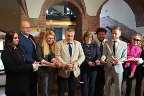 Pokaż zdjęcie: Grupa osób podczas uroczystego przecięcia biało-czerwonej wstęgi w zabytkowym wnętrzu z ceglanymi łukami. Uczestnicy stoją w półkolu, trzymają nożyczki, ubrani w formalne stroje, w tle winda ściany z cegły.
