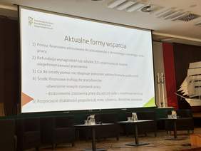 Pokaż zdjęcie: Na dużym telebimie wyświetla się prezentacja z logo PFRON. Tytuł slajdu „Aktualne formy wsparcia”