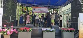 Pokaż zdjęcie: Scena plenerowa podczas festynu w Świdniku z okazji Dnia Godności Osób z Niepełnosprawnością Intelektualną. Na scenie grupa osób w strojach stylizowanych na dawne czasy, dekoracje kwiatowe i baner wydarzenia.