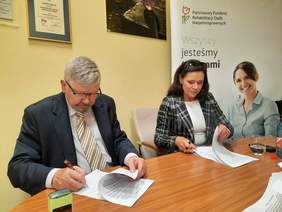 Pokaż zdjęcie: Przy stole siedzą dwie osoby: kobieta i mężczyzna. Podpisują dokumenty. Na stole leżą różne akta, pieczątki, telefony. 