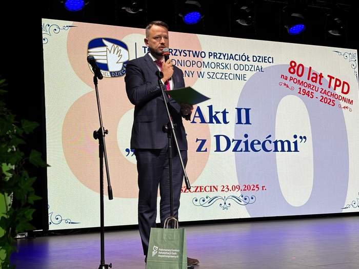 Pokaż zdjęcie: Mężczyzna w ciemnym garniturze i krawacie stoi na scenie, przemawiając do mikrofonu zamontowanego na stojaku. W lewej ręce trzyma złożoną zieloną kartę, a na scenie, tuż pod nim, leży zielona papierowa torba z logo Państwowego Funduszu Rehabilitacji Osób Niepełnosprawnych. W tle znajduje się duży, podświetlany ekran sceniczny, na którym wyświetlana jest grafika związana z obchodami jubileuszu. Tekst na ekranie, w języku polskim, informuje o uroczystości: "TOWARZYSTWO PRZYJACIÓŁ DZIECI ZACHODNIOPOMORSKI ODDZIAŁ REGIONALNY W SZCZECINIE", a duży, półprzezroczysty napis po prawej głosi "80 LAT TPD na POMORZU ZACHODNIM 1945 - 2025". Centralny tekst na ekranie to "Akt II z Dziećmi" oraz data "SZCZECIN 23.09.2025 r.".