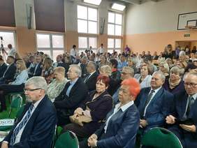 Pokaż zdjęcie: Na widowni siedzi tłum ludzi. Wszyscy patrzą na scenę. Są elegancko ubrani. Niektórzy mają ze sobą kwiaty lub upominki. 