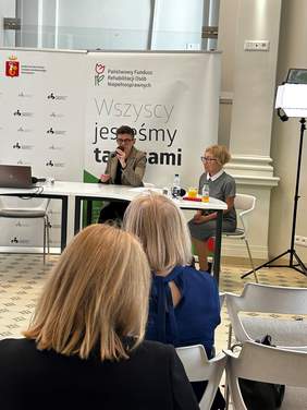 Pokaż zdjęcie: Dwie kobiety siedzą przy białym stole. Za nimi dwa rollupy z logo. Kobieta z lewej mówi przez mikrofon.