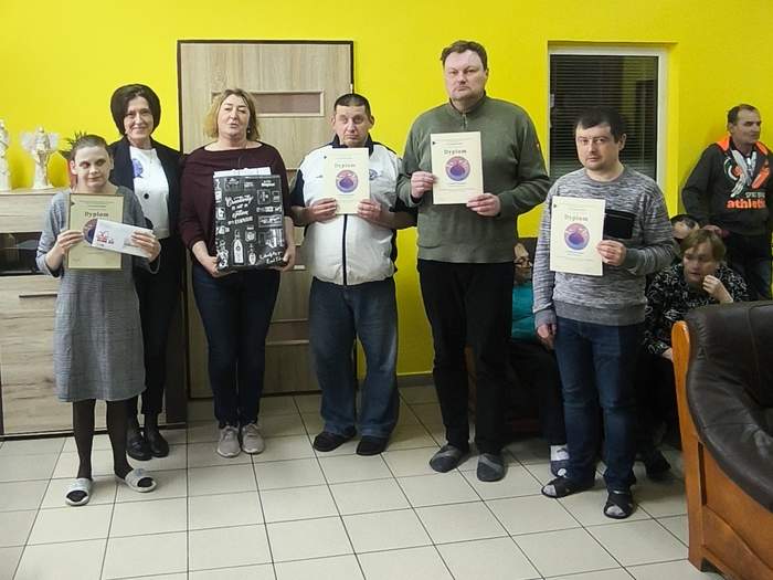 Pokaż zdjęcie: Grupa osób stojąca w jasnym pomieszczeniu na tle żółtej ściany, trzymająca dyplomy i prace plastyczne. W tle widoczne meble, gabloty z pucharami oraz kilka osób siedzących na kanapie.