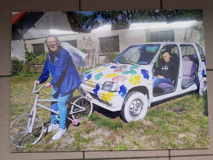 Pokaż zdjęcie: Fotografia przedstawiająca chłopaka w aucie i dziewczynę na rowerze, wszystko jest w różnych kolorach, jest słonecznie