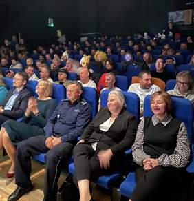 Widownia w kinie lub teatrze wypełniona ludźmi siedzącymi na niebieskich fotelach. W pierwszych rzędach siedzą dorośli, w tym mężczyzna w mundurze policjanta i kilka elegancko ubranych kobiet. Część osób na dalszych rzędach ma nietypowe nakrycia głowy lub charakteryzację.
