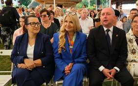 Pokaż zdjęcie: Osoby siedzące w pierwszym rzędzie podczas plenerowego wydarzenia. W tle liczna publiczność oraz drewniany budynek, uczestnicy obserwują program uroczystości.