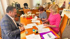 Pokaż zdjęcie: po lewej przy stole siedzi mężczyzna na przeciwko niego dwie kobiety wszyscy trzymają w dłoniach długopisy i podpisują dokumenty rozłożone na stole.