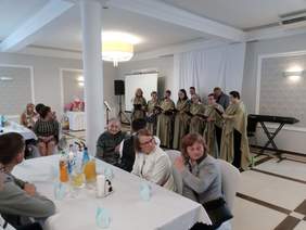 Pokaż zdjęcie: Na scenie stoi chór grupa siedmiu kobiet ubranych w takie same zielone długie tuniki, na pierwszym planie widać gości siedzących przy stołach