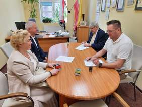 Pokaż zdjęcie: Przy podłużnym stole siedzi kobieta i trzech mężczyzn. Podpisują umowę. Na stole leżą różne dokumenty i pieczątki. 