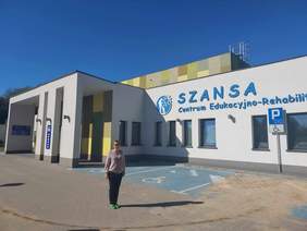 Pokaż zdjęcie: Przed budynkiem, na którym jest napisane Centrum Edukacyjno-Rehabilitacyjne „Szansa”, stoi kobieta. Widać też miejsca parkingowe. Dzień jest słoneczny. 