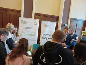 Pokaż zdjęcie: Kobieta i mężczyzna reprezentujący Oddział Wielkopolski PFRON udzielają informacji uczestnikom Giełdy Pracy dla Osób z Niepełnosprawnością. W tle znajdują się dwa banery: PFRON oraz CIDON.