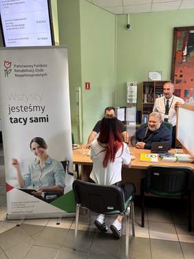 Pokaż zdjęcie: Kobieta stoi przy stoliku naprzeciw dwóch siedzących mężczyzn. Na stole leżą dokumenty i materiały informacyjne, a w tle widoczny jest kolejny mężczyzna stojący obok stanowiska.