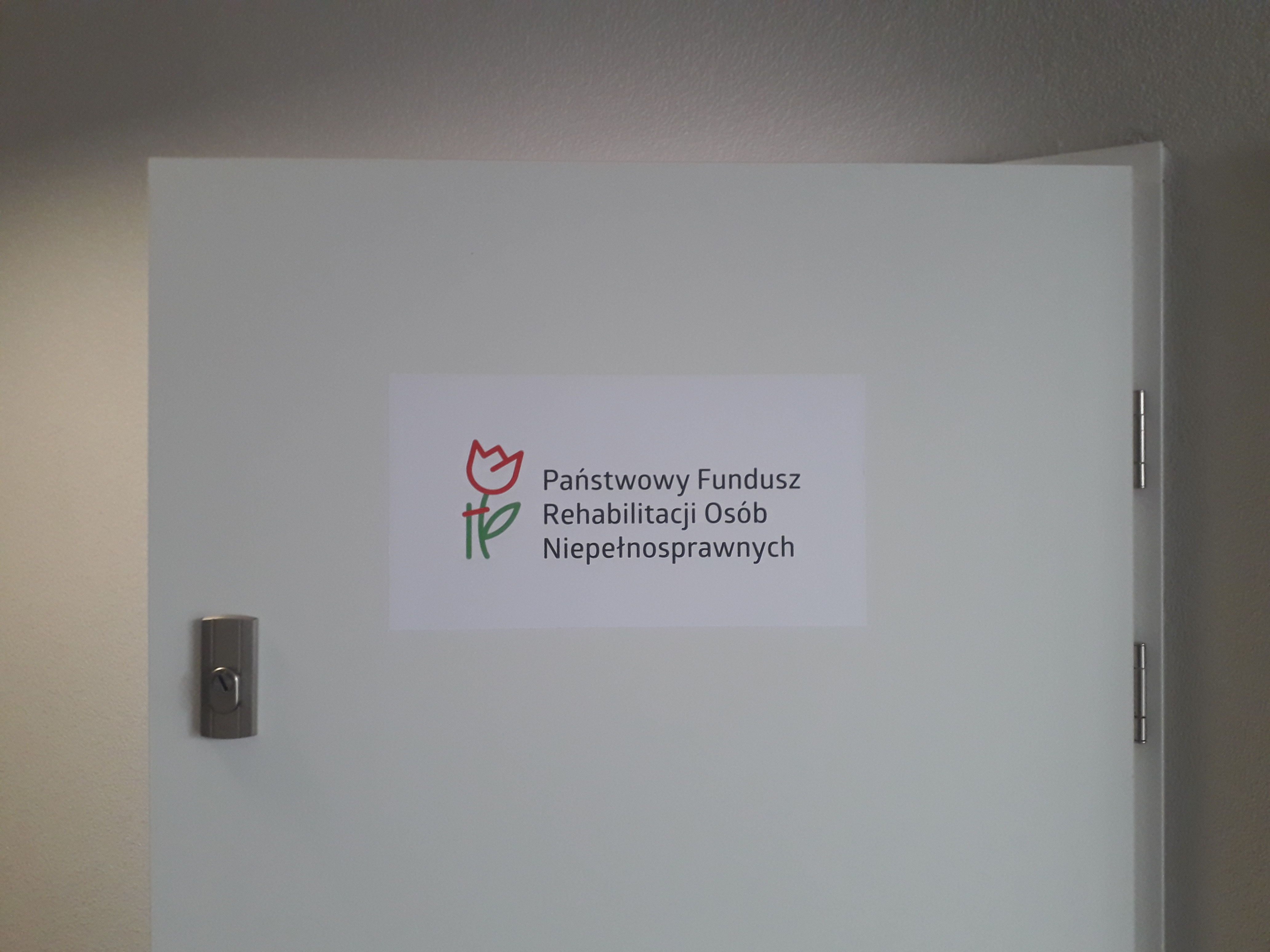 Wejście do Oddziału Dolnośląskiego Białe drzwi na tle jasnej ściany. Na drzwiach centralnie umieszczona tablica z logo PFRON