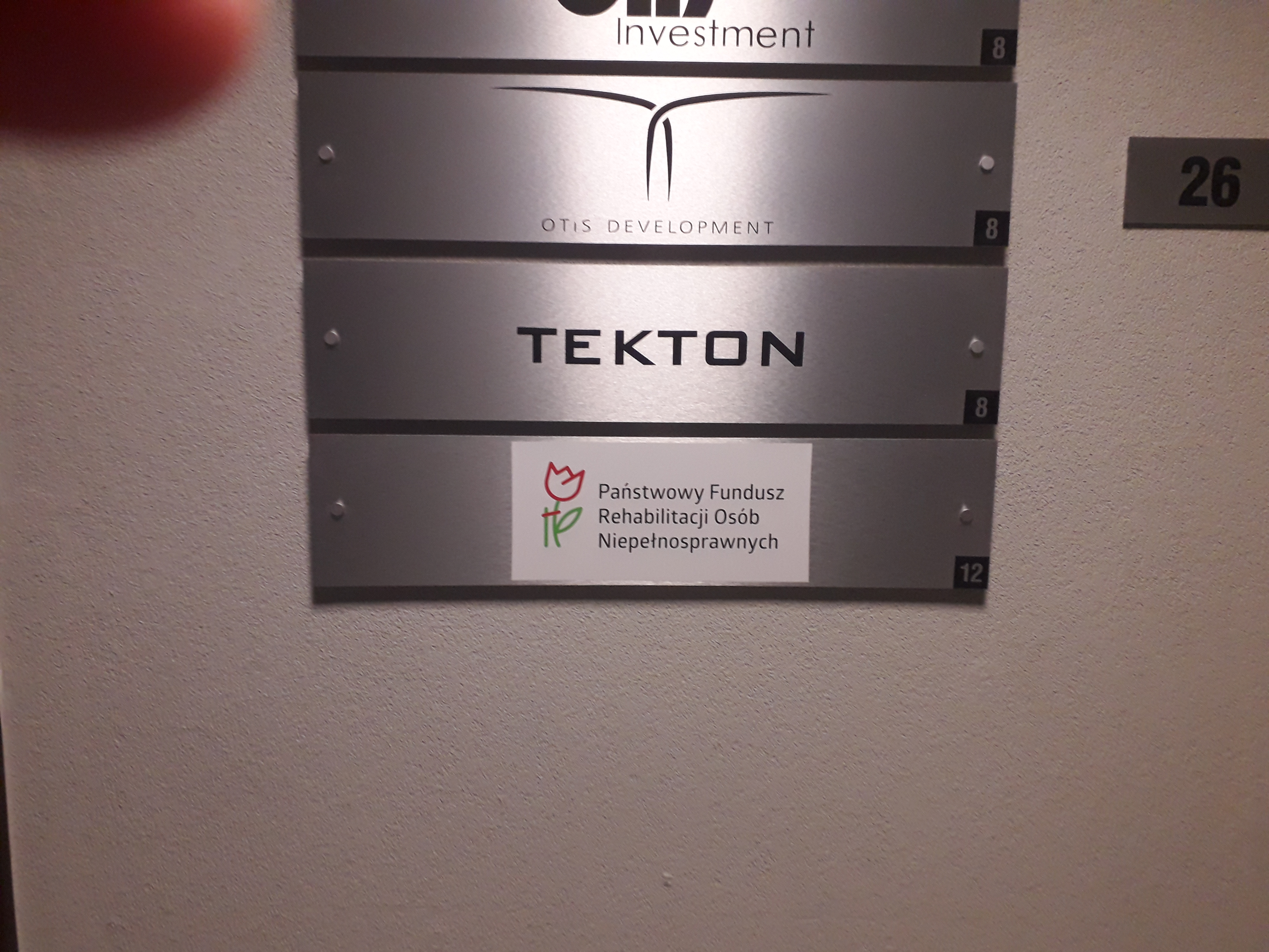 Tablica informacyjna Tablica informacyjna zawieszona pośrodku jasnej ściany. Na srebrnym tle umieszczono poziomo nazwy firm. Na ostatniej pozycji, pośrodku tablicy biały prostokąt, na którym umieszczono logo PFRON. Po prawej stronie od tablicy widoczny czarny numer 26 na srebrnym tle, przyczepiony na ścianie