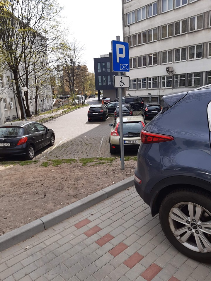 Widok na parking z miejscem dla Osoby z niepełnosprawnością. Widok placu parkingowego otoczonego budynkami. po prawej stronie budynek o jasnej elewacji, trzykondygnacyjny, za nim dwukondygnacyjny budynek o ciemnoszarej elewacji z podcieniami. Po lewej stronie budynek o 3 kondygnacjach. Przed budynkiem tym rosną drzewa. Na placu parkingowym samochody, na pierwszym planie granatowy samochód, który stoi na miejscu parkingowym wyznaczonym dla osoby niepełnosprawnej. Miejsce oznaczone znakiem pionowym.