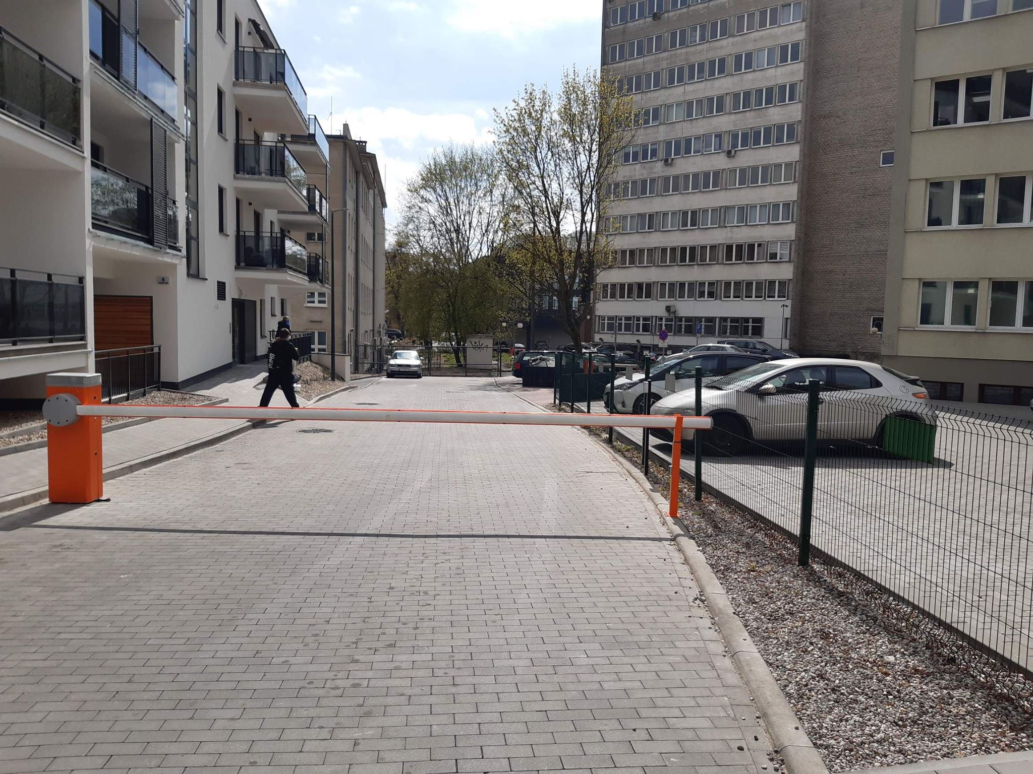 Wjazd na parking. Ulica, po lewej stronie mamy budynek mieszkalny, jasno-biały z balkonami. Pośrodku ulicy szlaban. Ulica prowadzi do jasnego budynku o kilku kondygnacjach, przed którym rosną wysokie drzewa. po prawej stronie od szlabanu widzimy ogrodzony parking, na którym zatrzymały się samochody
