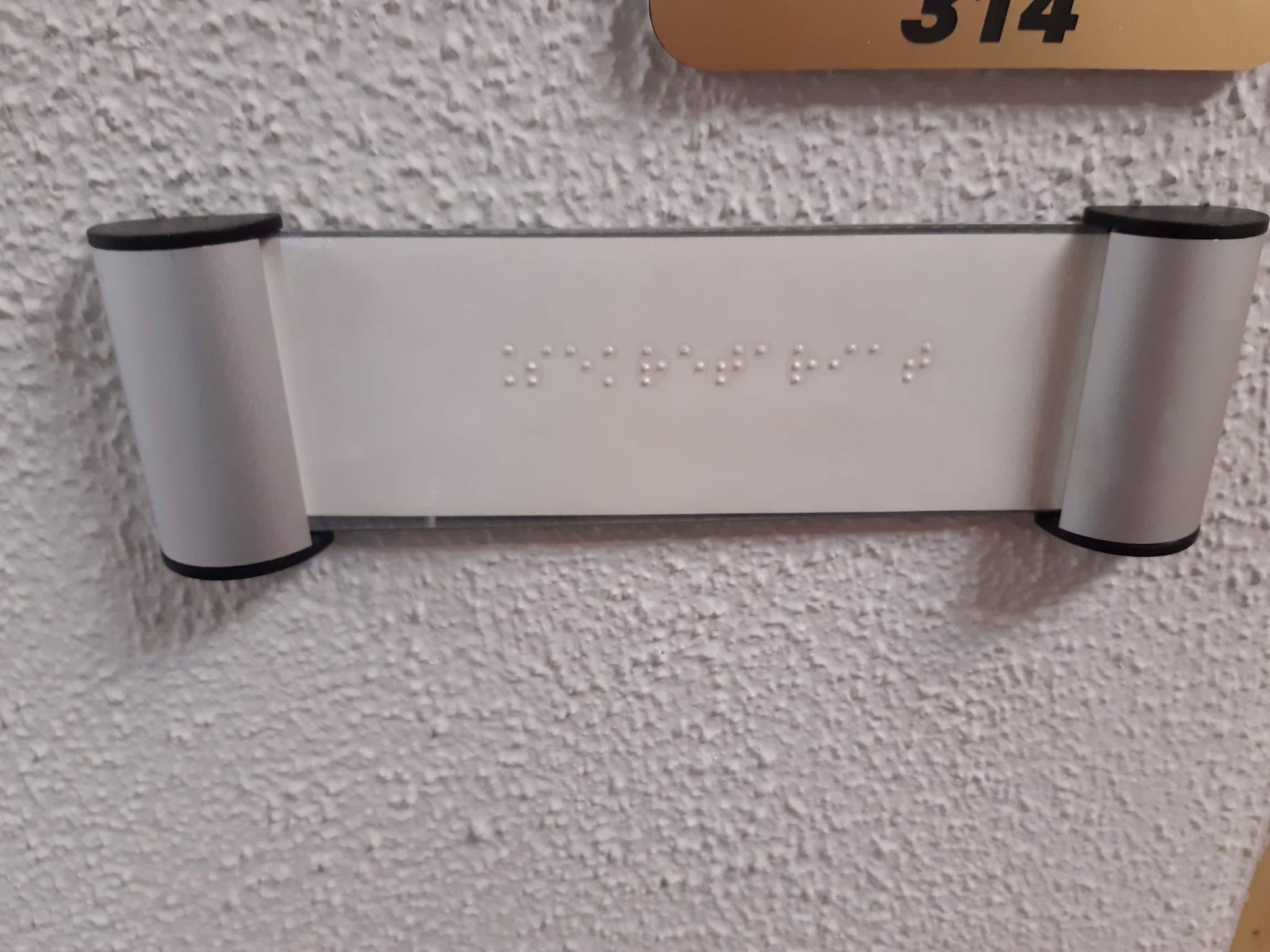 Tabliczka informacyjna w alfabecie Braille'a Na jasnej ścianie jasnoszara tabliczka z napisem w języku Braille’a.