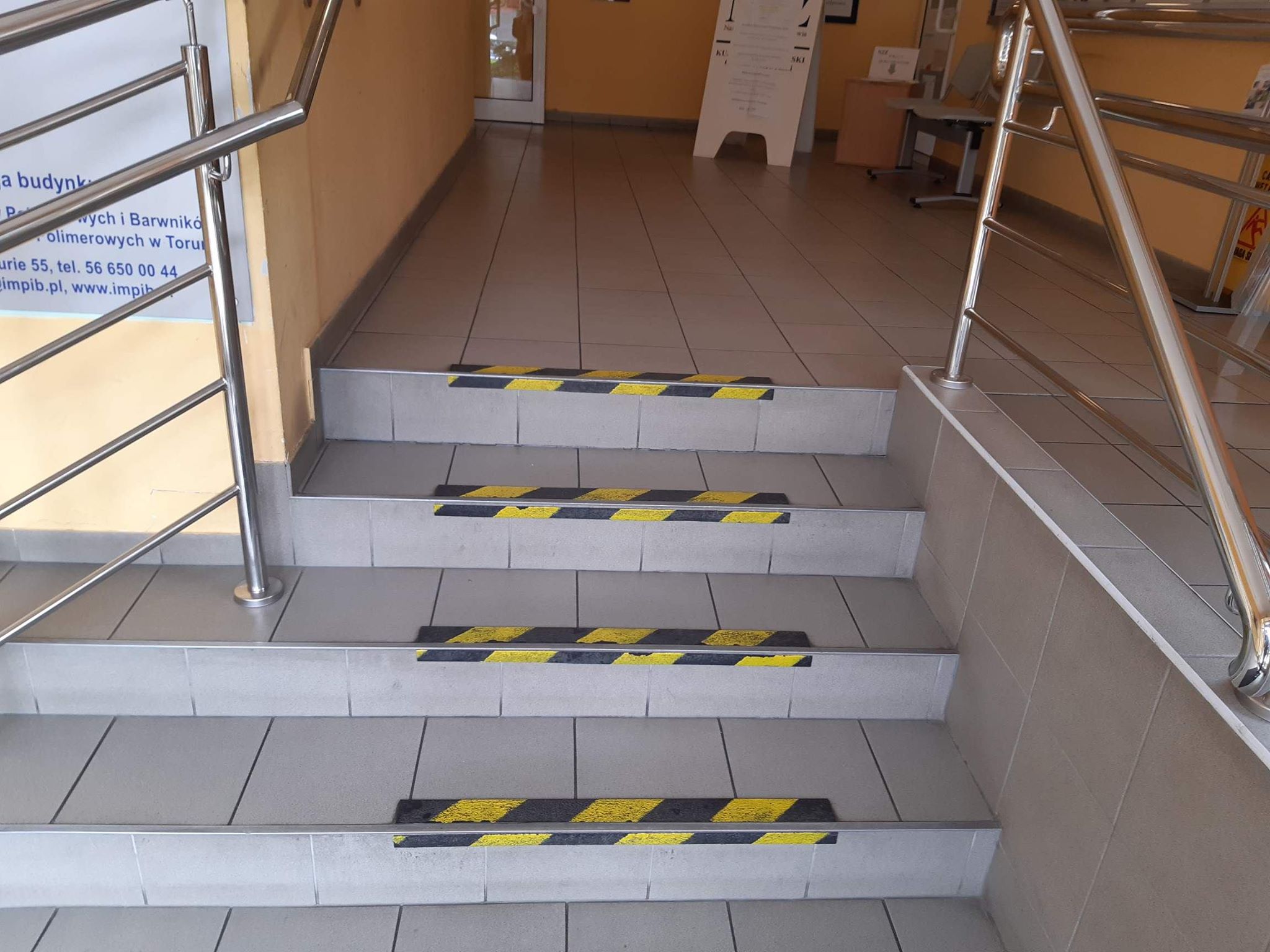 Schody wewnętrzne. Schody obłożone jasnoszarymi płytkami, na krawędziach schodów czarno-żółte taśmy mające skontrastować stopnie schodów. Po lewej i po prawej stronie widzimy poręcze wykonane ze stali nierdzewnej. Po lewej stronie za poręczą widać tablicę informacyjną. Schody prowadzą na poziom, gdzie widzimy białą tablicę informacyjną i drzwi po lewej stronie.