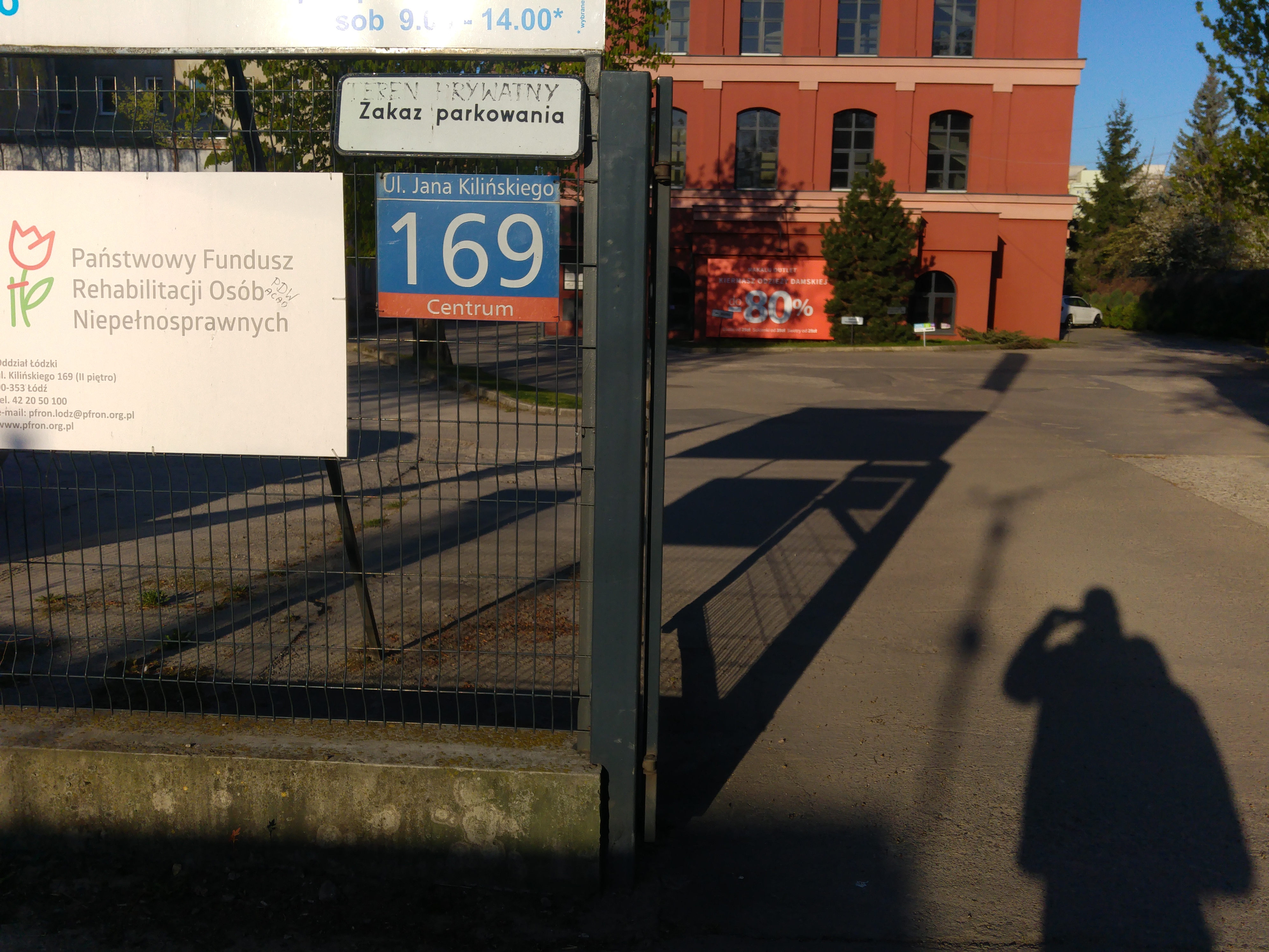 Wjazd na dziedziniec. Ogrodzenie z drucianej siatki na betonowej podbudowie. Na ogrodzeniu duża tablica z logiem i napisem „Państwowy fundusz Rehabilitacji Osób Niepełnosprawnych”. Na prawo tabliczka adresowa „ulica Jana Kilińskiego 169, dzielnica centrum”. Nad tabliczką adresową tabliczka zakaz parkowania teren prywatny. Po prawej stronie otwarta brama prowadząca na dziedziniec dużego budynku. Widzimy tylko 3 kondygnacje, ceglany kolor ścian z jasno ceglanymi gzymsami. Na każdej kondygnacji widzimy dosyć duże okna. Przed budynkiem rosną drzewa, pomiędzy którymi umieszczona jest tablica z dużym napisem „-80%”