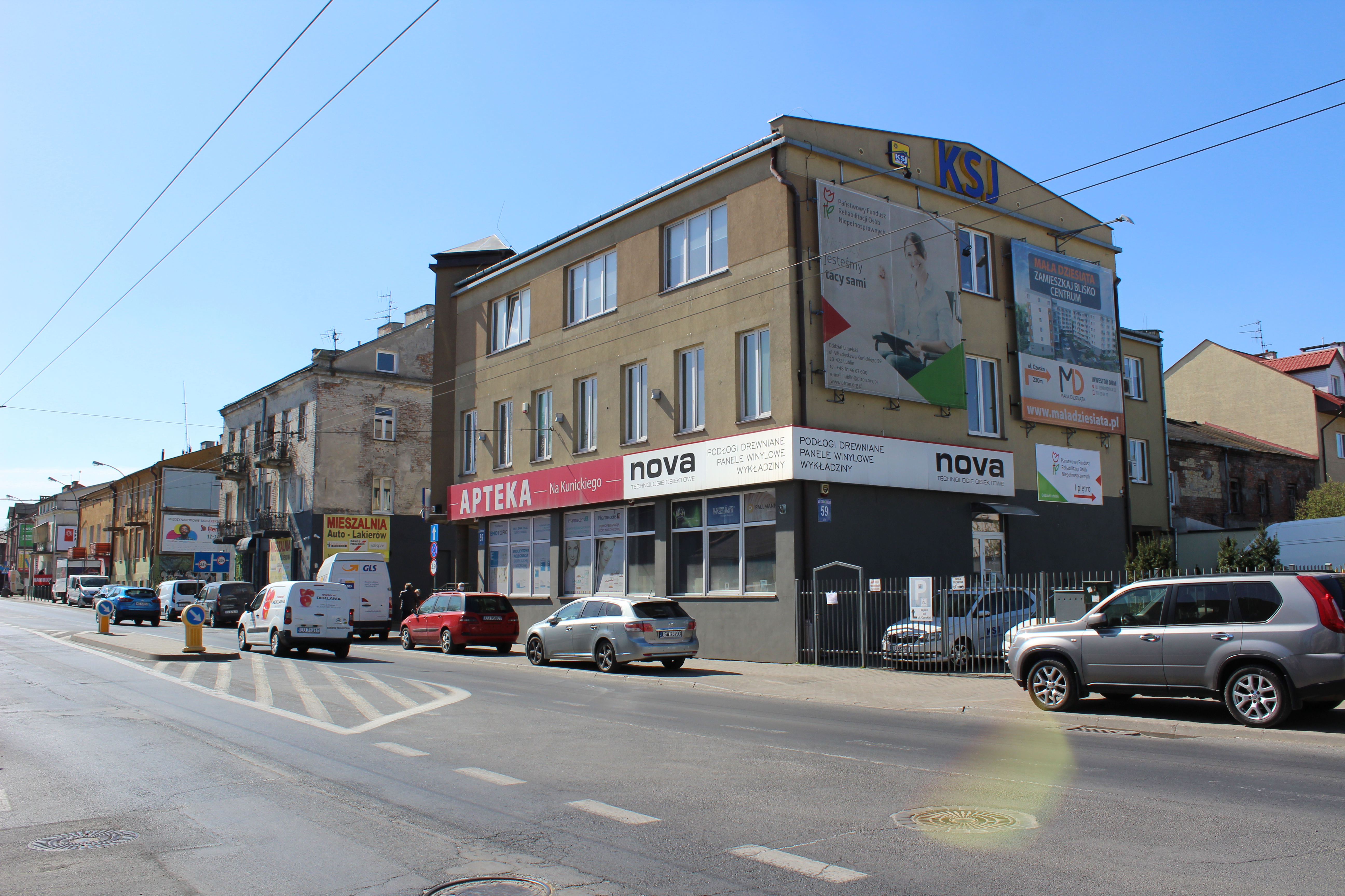 Ulica, na chodniku zaparkowane samochody. Po prawej stronie stalowe ogrodzenie z furtką, prowadzącą na dziedziniec budynku. Budynek dwukolorowy. Parter w ciemnoszarym kolorze, pozostałe kondygnacje w ciemnożółtym. Od strony ulicy liczne okna. na ścianie od dziedzińca widać duży baner reklamowy, a pod nim baner PFRON. Granicę między parterem, a wyższymi kondygnacjami podkreślają plansz reklamowe z napisem "apteka"