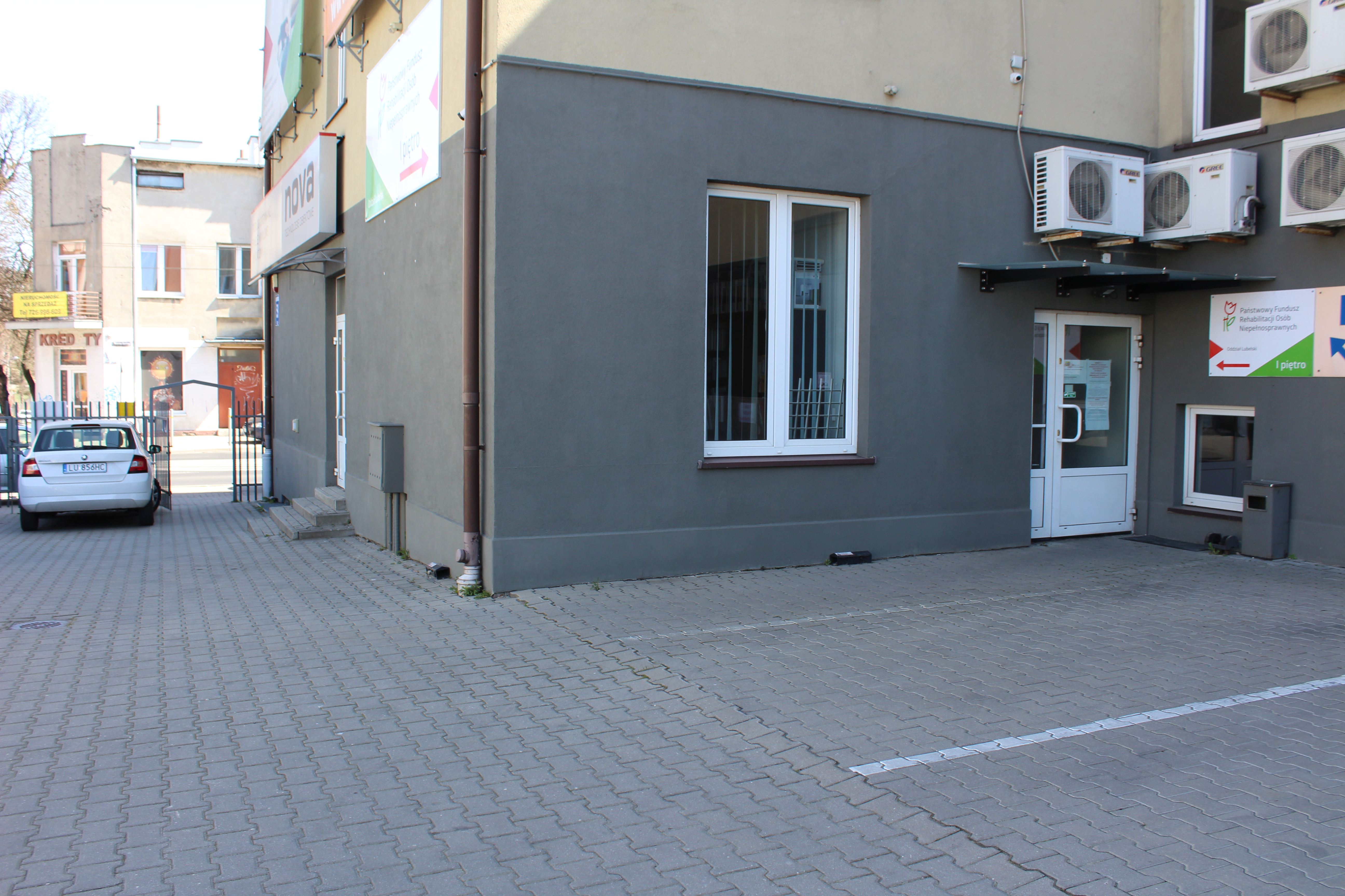 Plac z kostki brukowej z zaznaczonymi liniami parkingowymi, po prawej stronie budynek z kondygnacją parteru pomalowaną na szaro, z dużym oknem. Po prawej storni od okna widać drzwi wejściowe do budynku. Nad drzwiami daszek ochronny, a ponad nim klimatyzatory, po których prawej stronie zawieszone logo PFRON. Po lewej storni od budynku widać zaparkowany samochód, a w jego tle otwartą furtkę w ogrodzeniu.
