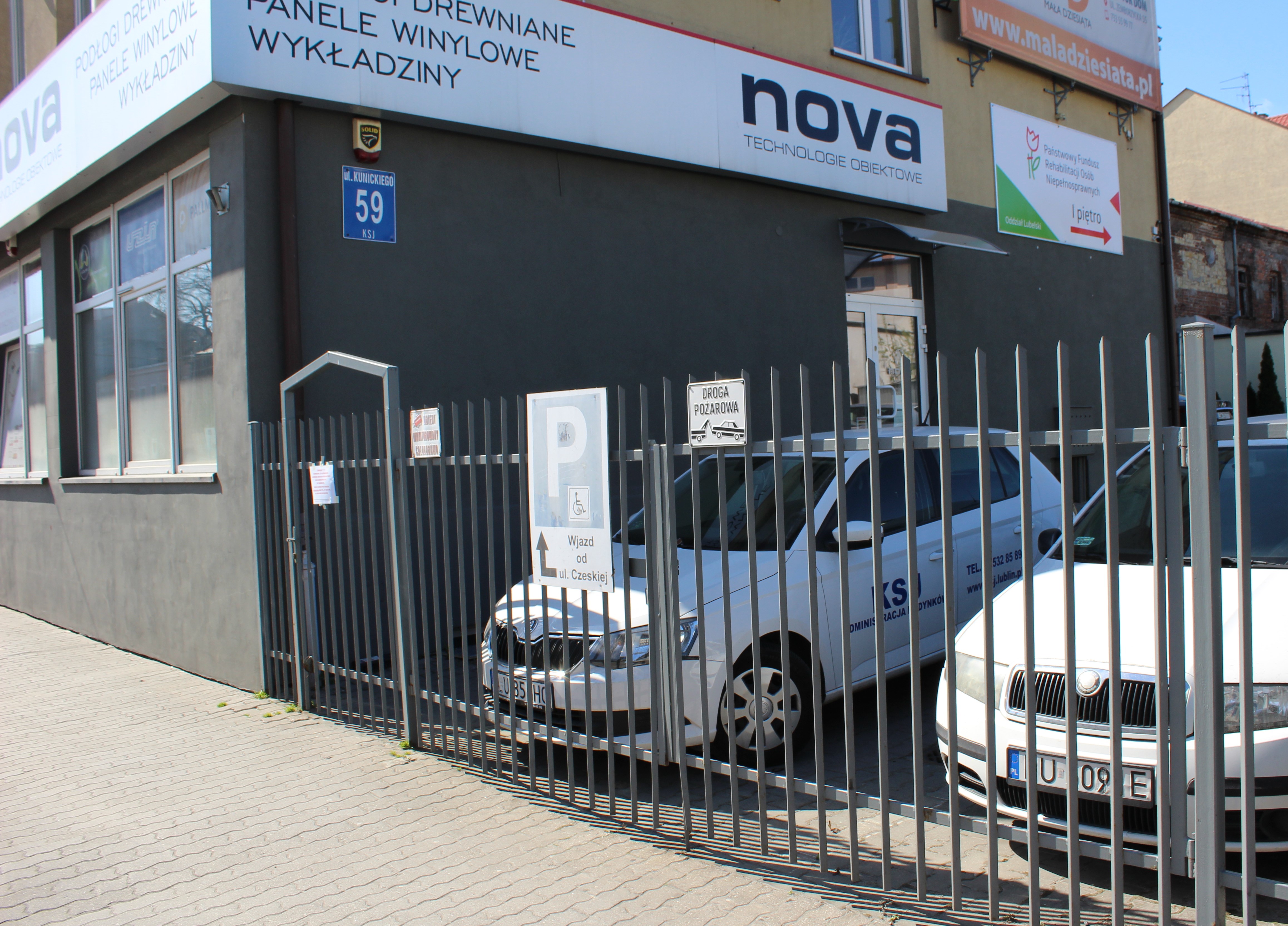 W tle budynek o szarej elewacji, na lewej scianie rząd okien. Poprawej stronie widać metalowe ogrodzenie z furtką. Po prawej stronie od furtki widać znak parkingu z piktogramem osoby niepełnosprawnej. Za ogrodzeniem widać zaparkowane samochody. 