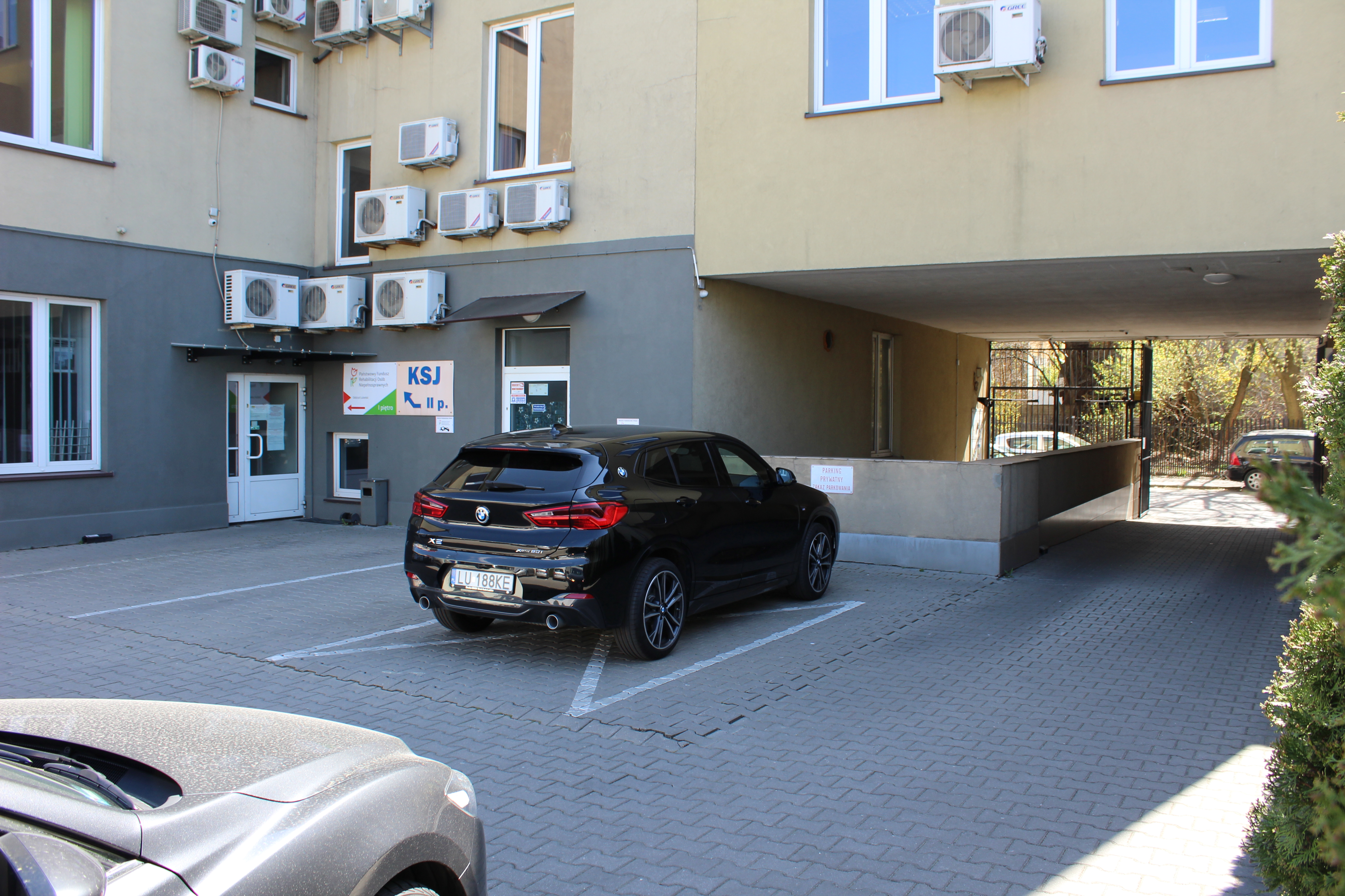 W lewym dolnym rogu widać fragment zaparkowanego samochodu. Po lewej stronie widać wyjazd z parkingu na ulicę przez bramę. Na lewo od bramy widać centralnie zaparkowany samochód. Za samochodem widać częściowo zasłonięte wejście do budynku, po którego lewej stronie widać na ścianie logo PFRON. W rogu budynku następne drzwi wejściowe, nad którymi zawszone na ścianie liczne klimatyzatory