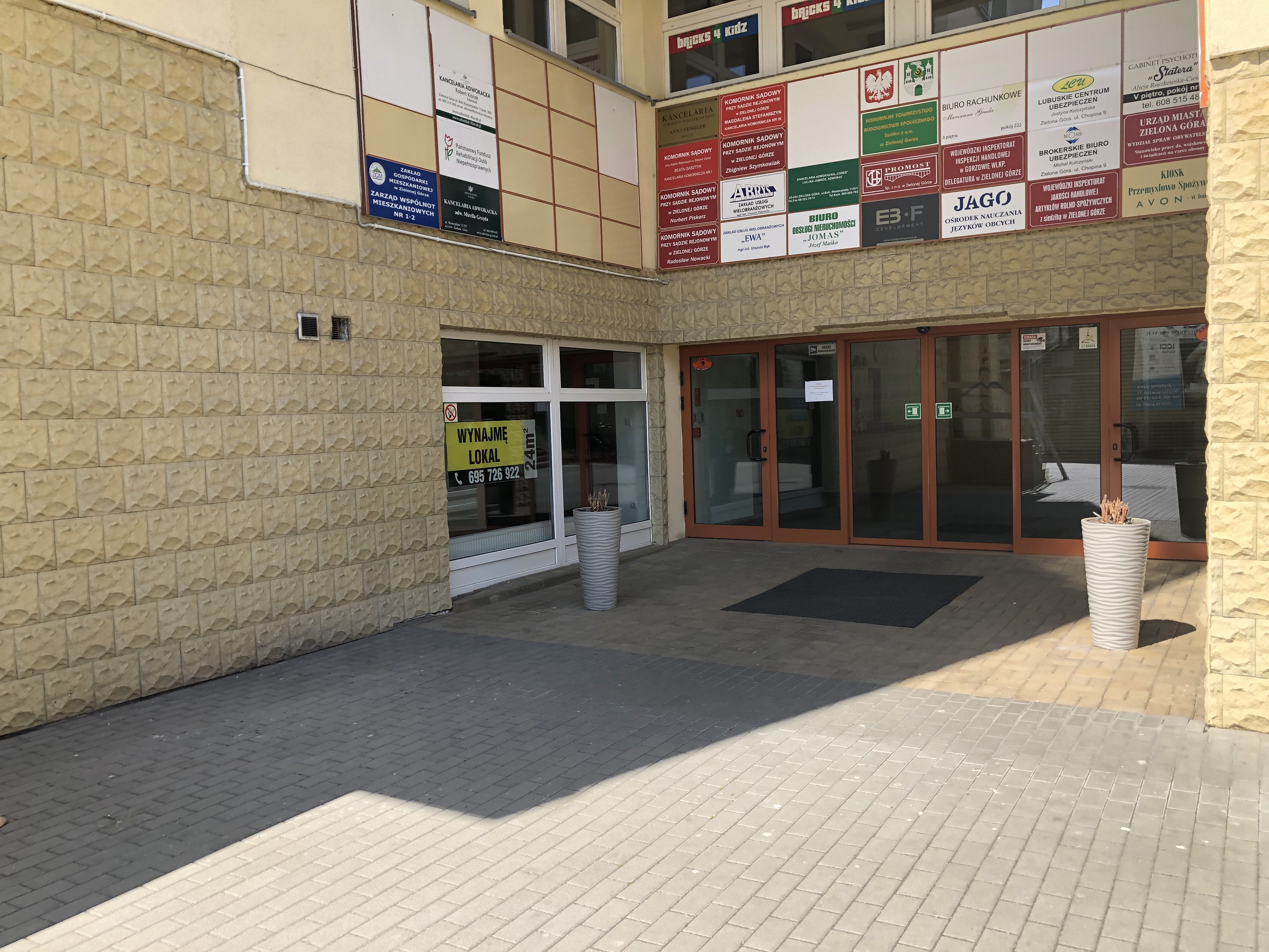 Wejście do budynku. Wejście do budynku. W tle widać brązowe drzwi automatyczne, rozsuwane, na nimi mnóstwo tablic z nazwami instytucji, które mieszczą się w budynku. Pomiędzy nimi znajduje się tablica z logo PFRON. Na lewo od wejścia duże okna wykonane w białym kolorze. Ściana obłożona kamieniem w kolorze piaskowca - ciemnożółtym.