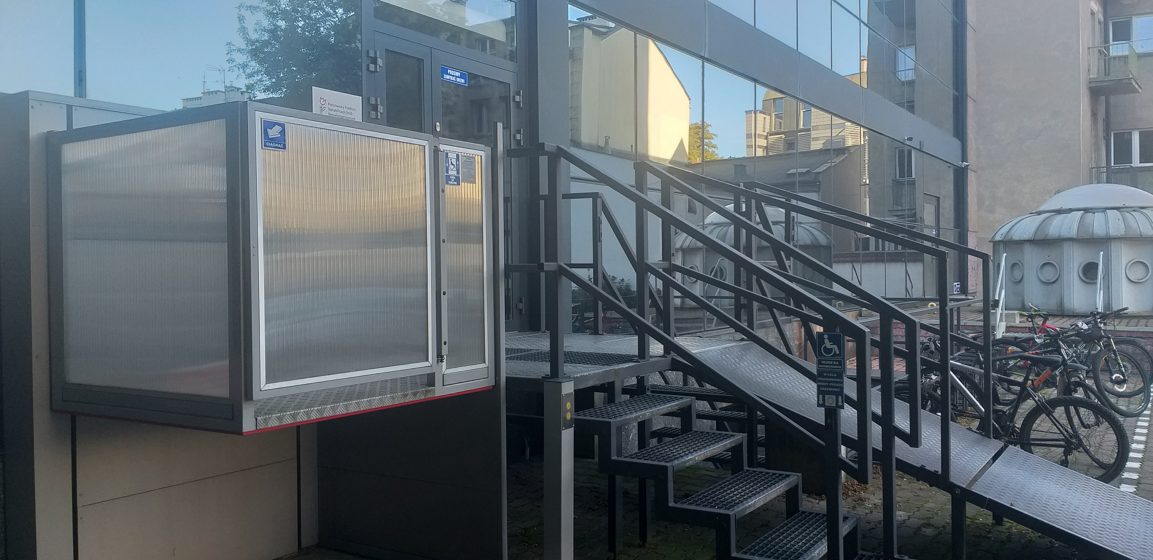 Widok windy dla wózków - ujęcie drugie Widok na windę dla wózków. Po prawej stronie od windy, na poziomie ziemi słupek, gdzie są umieszczone przyciski do sterowania windą. Za windą widać podest, do którego prowadzą ażurowe schody i rozkładana pochylnia. Na prawo od pochylni zaparkowane rowery, w tle przeszklona ściana budynku