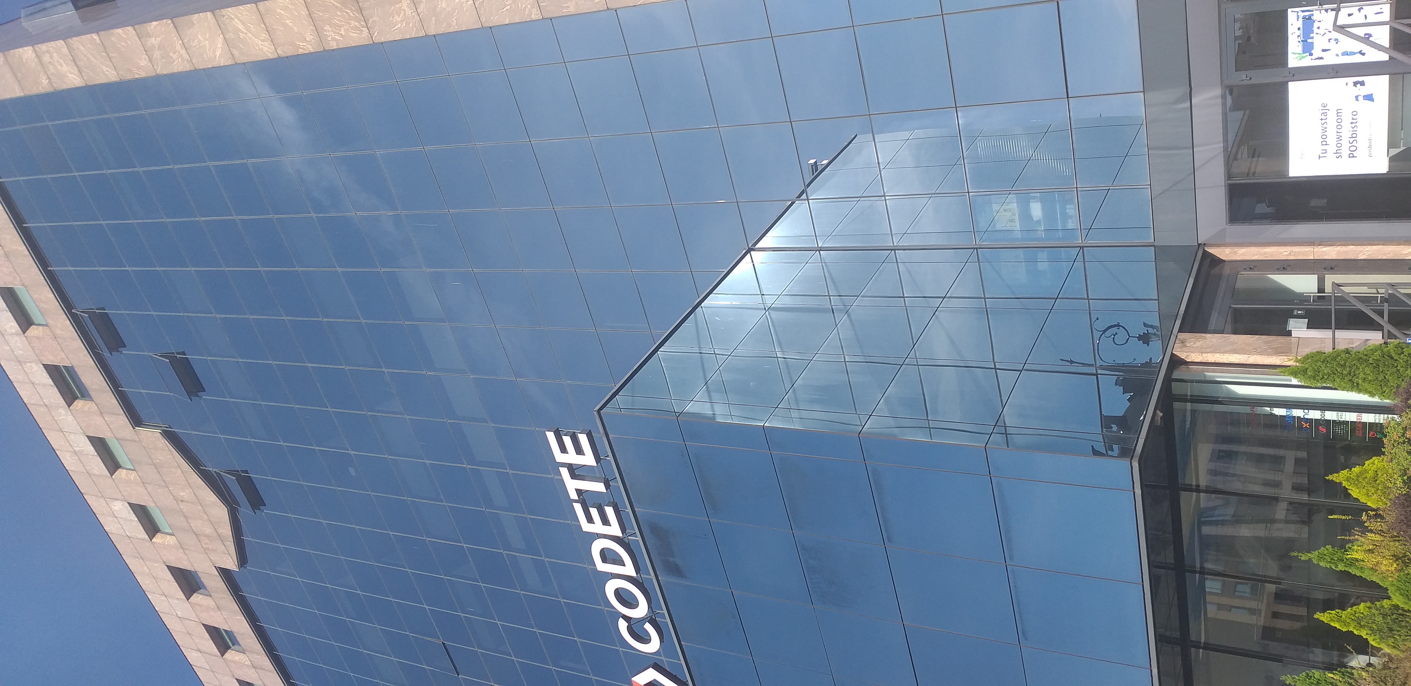 Siedziba Oddziału Małopolskiego PFRON Budynek biurowy ze szklaną ścianą, w której odbija się bezchmurne niebo. Na pierwszym planie niższa część budynku, na której dachu umieszczono napis „Codete”. W prawym, dolnym rogu widać drzwi wejściowe do budynku.
