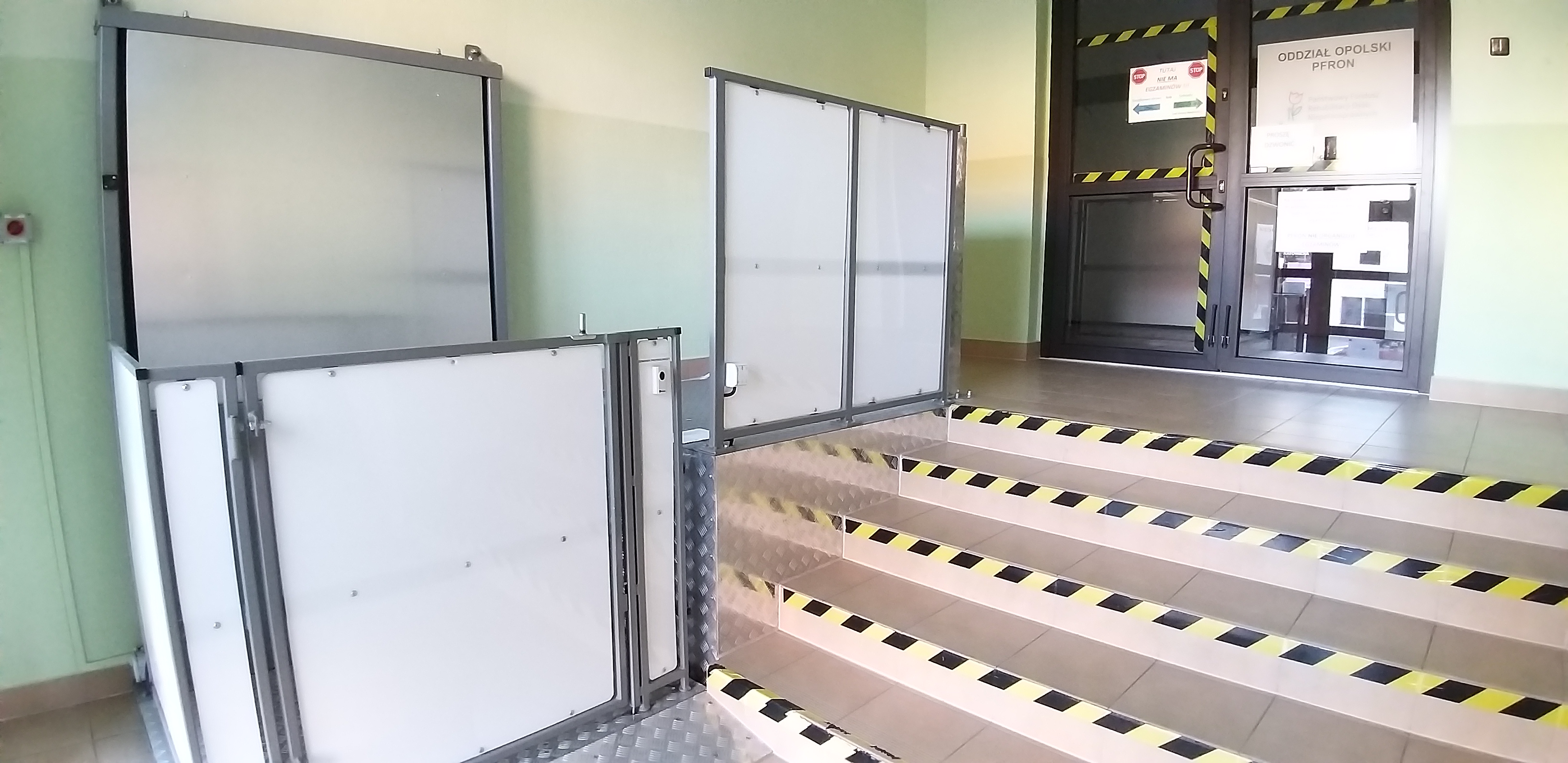 Schody wejściowe do Oddziału Opolskiego PFRON Schody wyłożone jasno-brązowymi płytkami. Na krawędziach schodów taśmy ostrzegające. Po lewej stronie od schodów konstrukcja windy dla wózków, otoczona białymi ściankami. Nad schodami widać drzwi wejściowe skontrastowane pasami czarno żółtymi z dużym napisem „Oddział Opolski PFRON”
