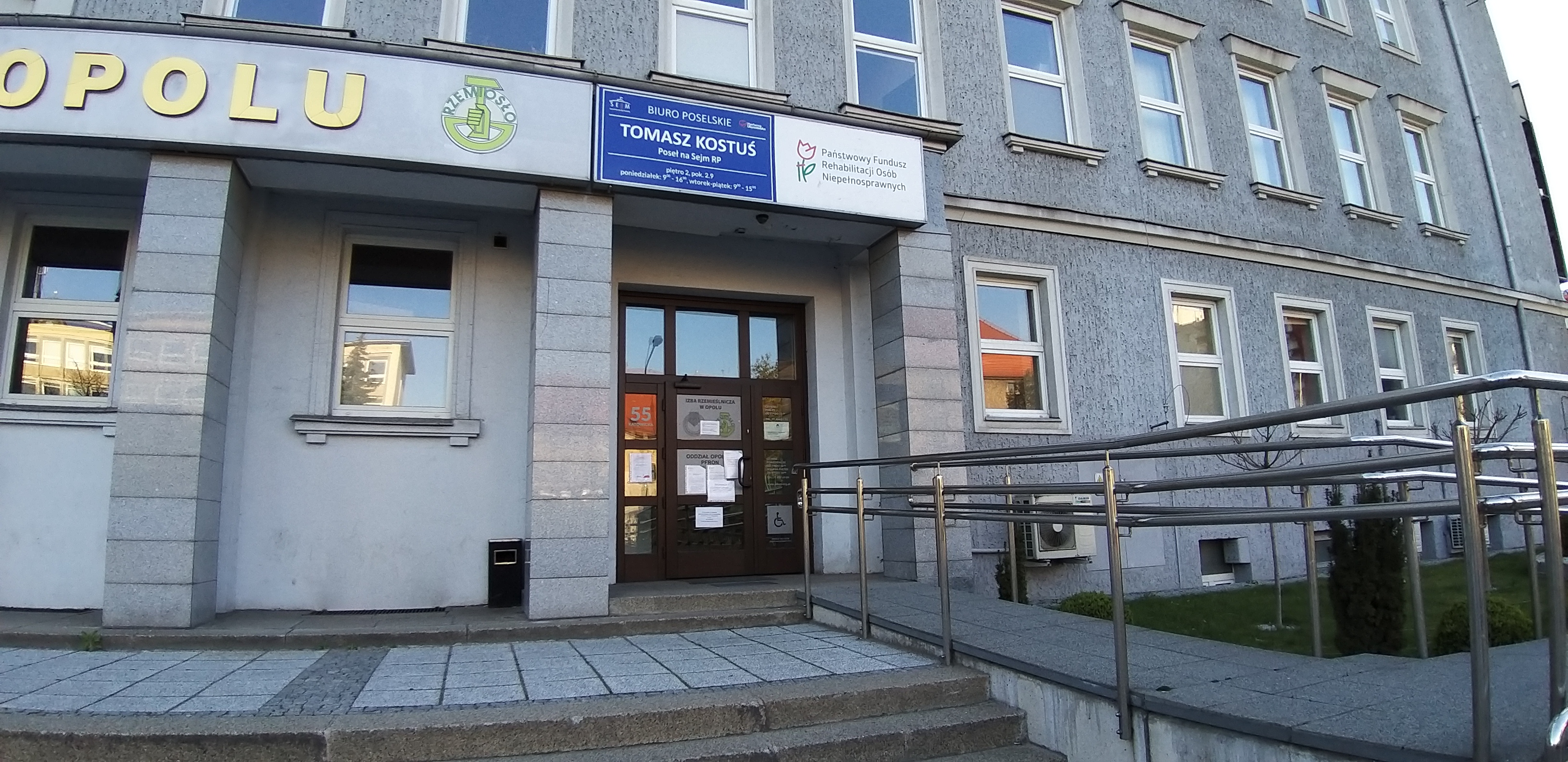 Siedziba Oddziału Opolskiego PFRON - widok pochylni Jasno szary budynek biurowy, na pierwszym planie pochylnia dla wózków oraz schody prowadzące do drzwi wejściowych. Nad drzwiami, na gzymsie duża tablica z logo i napisem „Państwowy Fundusz Rehabilitacji Osób Niepełnosprawnych”; na lewo od tej tablicy, tablica niebieska z napisem „Tomasz Kostuś, poseł na Sejm RP”