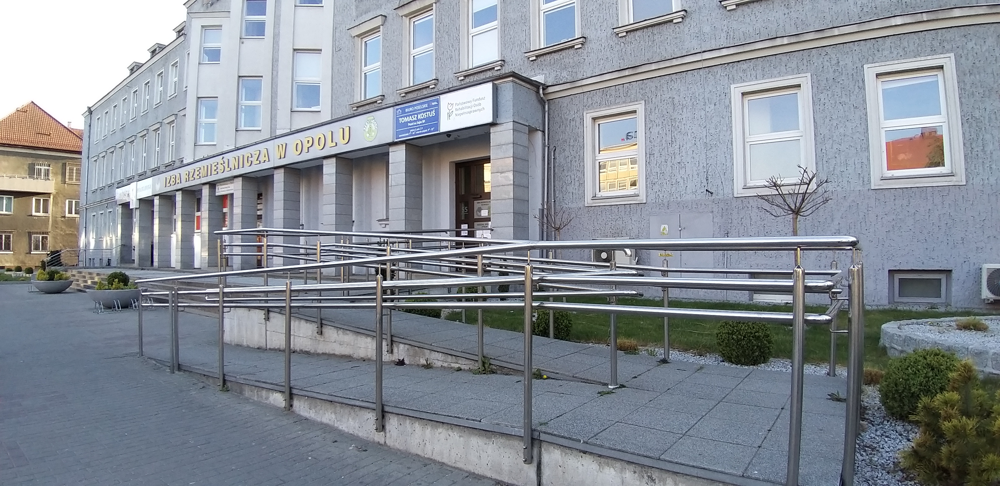 Wejście główne do siedziby Oddziału Opolskiego PFRON Jasno szary trzykondygnacyjny budynek biurowy z dużą ilością okien. Na pierwszym planie dwuzabiegowa pochylnia prowadząca do drzwi wejściowych. Nad głównym wejściem napis „Izba Rzemieślnicza w Opolu” oraz 2 tablice informacyjne, jedna z tekstem „Tomasz Kostuś, poseł na Sejm RP”, a druga biała z logo i napisem „Państwowy Fundusz Rehabilitacji Osób Niepełnosprawnych"